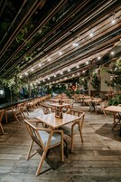 Un restaurante con mesas y sillas de madera, una zona de bar y un techo con vigas a la vista y luces colgantes.