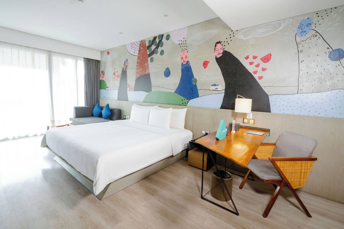 A hotel room with a large bed, a wooden desk, a chair, and a wall mural.