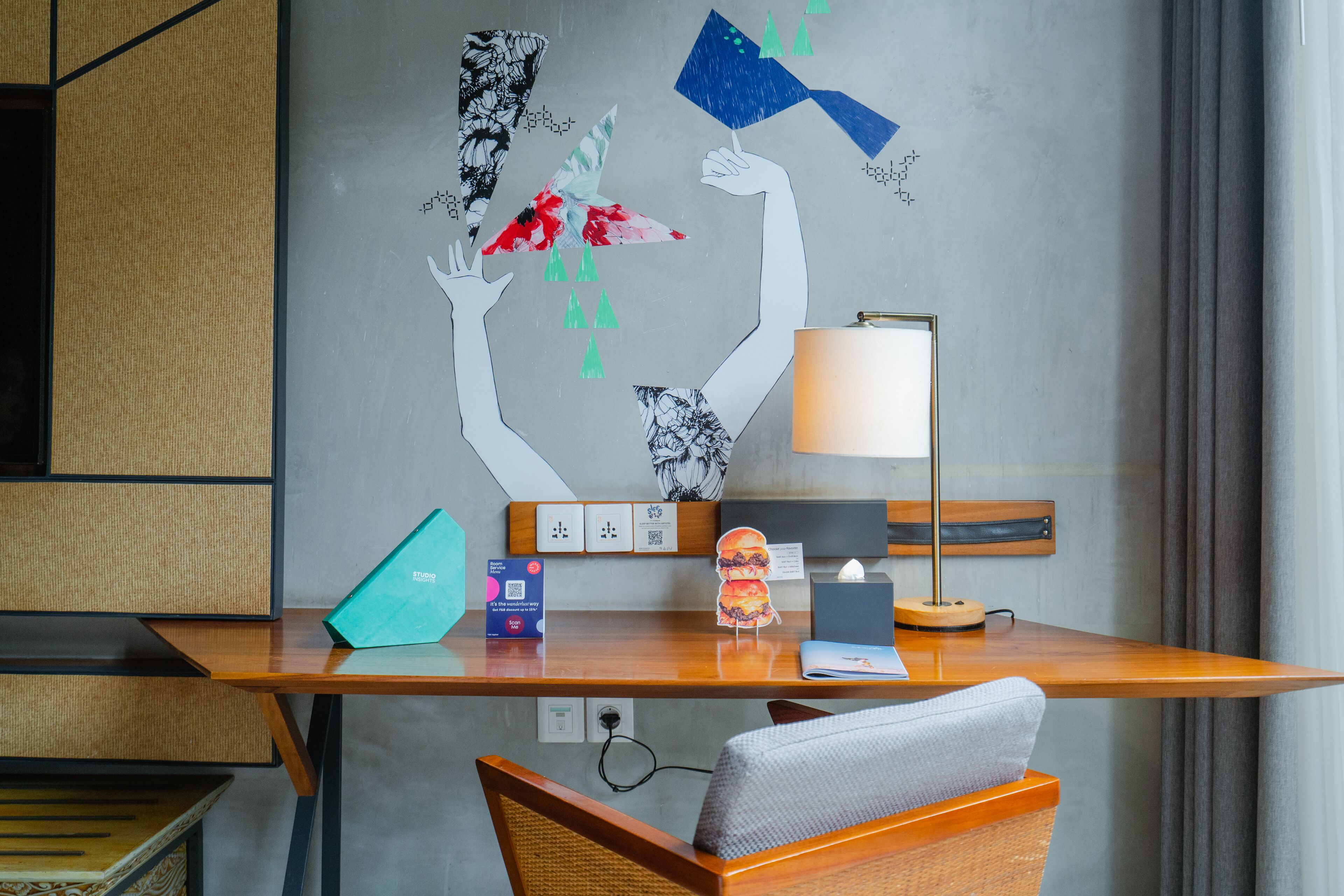 Una habitación de hotel moderna con un escritorio de madera, una lámpara, una silla y una pared decorada con arte abstracto.