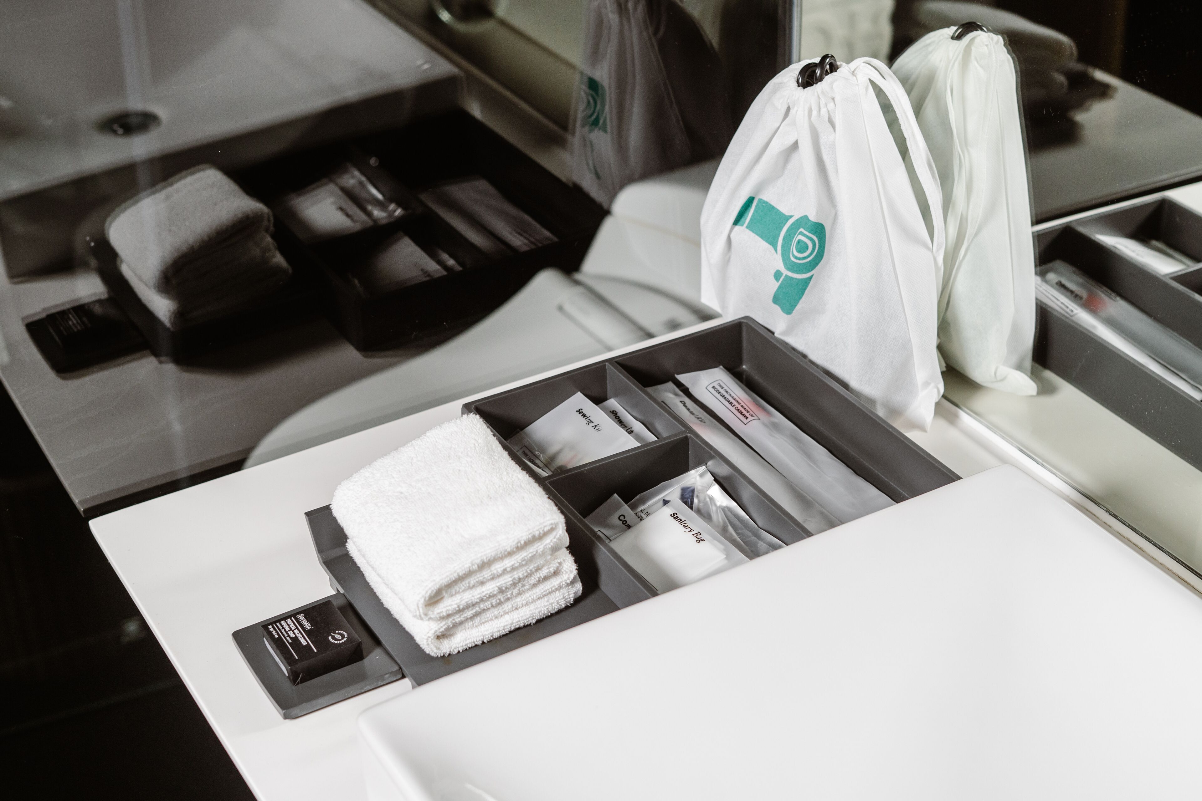 A neatly organized hotel room with a bed, a small table with a towel and toiletries, and a white bag with a logo.