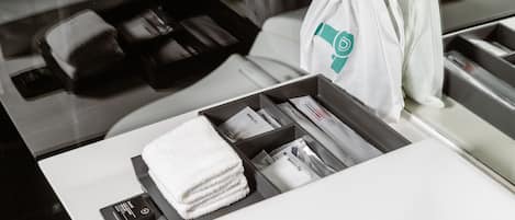 A neatly organized hotel room with a bed, a small table with a towel and toiletries, and a white bag with a logo.
