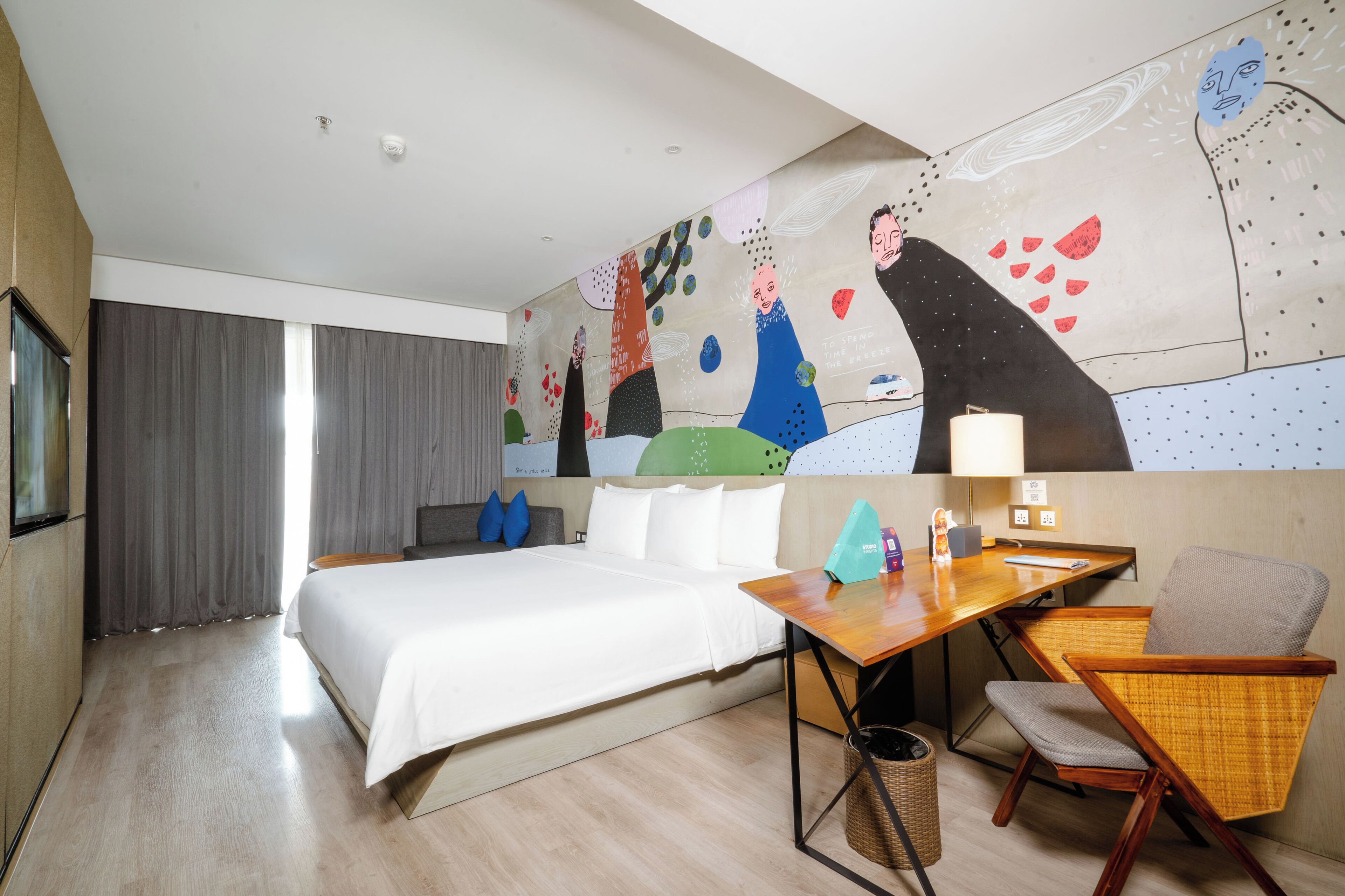 A hotel room with a large bed, a desk with a chair, a wall mural, and a window with curtains.