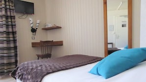 Un dormitorio con cama, una silla, una estantería con objetos y un televisor en la pared.