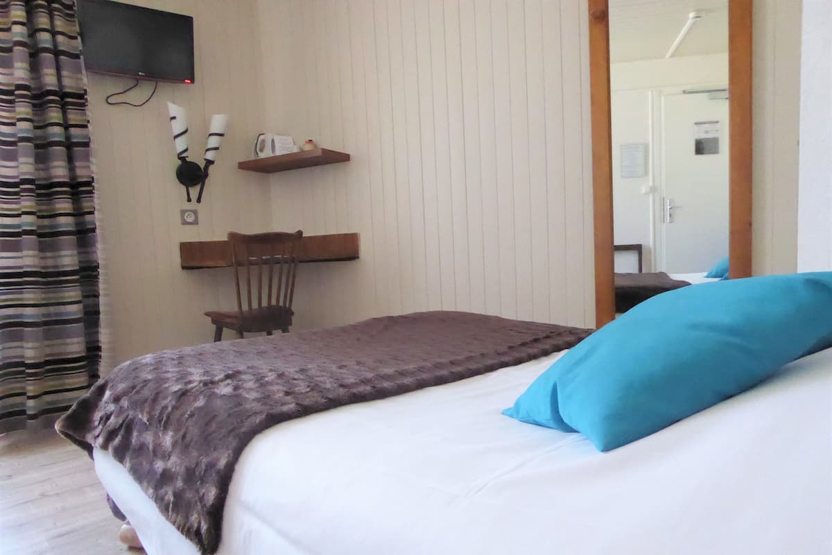 Un dormitorio con cama, una silla, una estantería con objetos y un televisor en la pared.