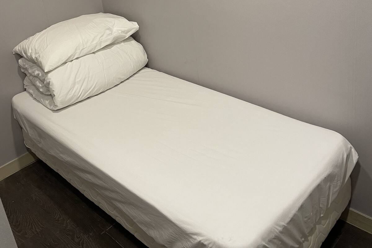 Une petite chambre avec un lit simple, un matelas et des oreillers blancs unis, un plancher en bois foncé et des murs de couleur gris clair.