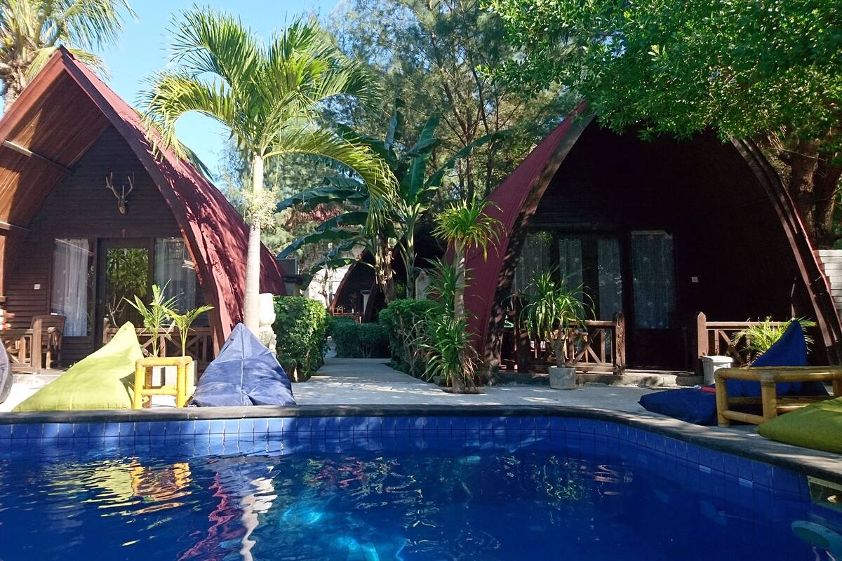 Zona de piscina amb dues cabanes de fusta, envoltada de plantes tropicals i una piscina ampla i blava.