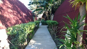 A narrow concrete pathway flanked by dense green foliage and a red corrugated metal wall. - Coral Beach 2 Bungalow (Gili Trawangan)