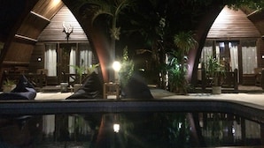 A nighttime view of a resort with two wooden-roofed cabins, a swimming pool, and surrounding palm trees. - Coral Beach 2 Bungalow (Gili Trawangan)