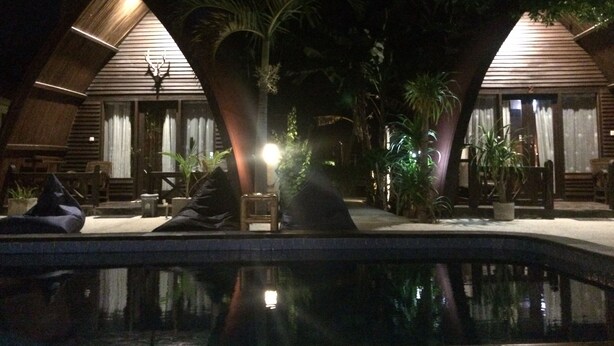 A nighttime view of a resort with two wooden-roofed cabins, a swimming pool, and surrounding palm trees.