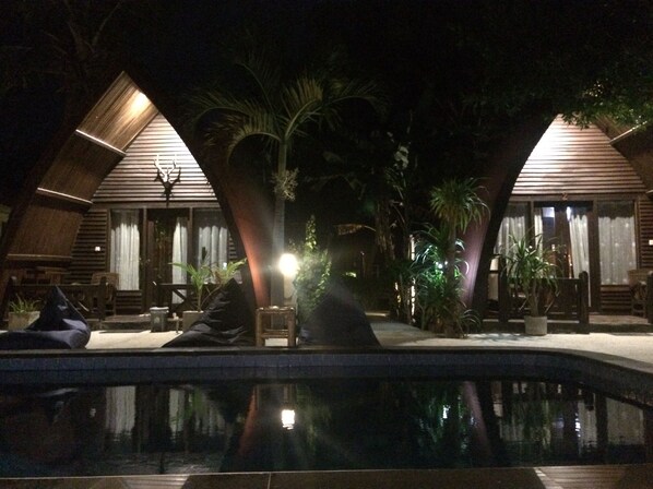 A nighttime view of a resort with two wooden-roofed cabins, a swimming pool, and surrounding palm trees. - Coral Beach 2 Bungalow (Gili Trawangan)
