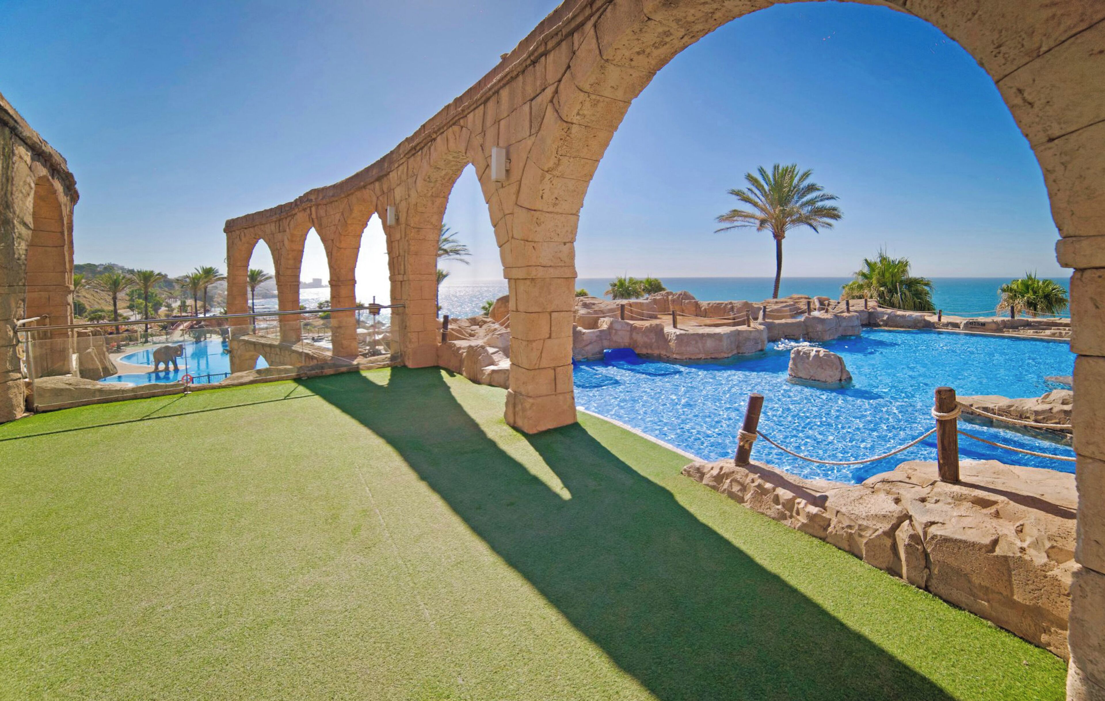 Une piscine extérieure bordée d’arches en pierre, un ciel dégagé et une vue sur la mer.