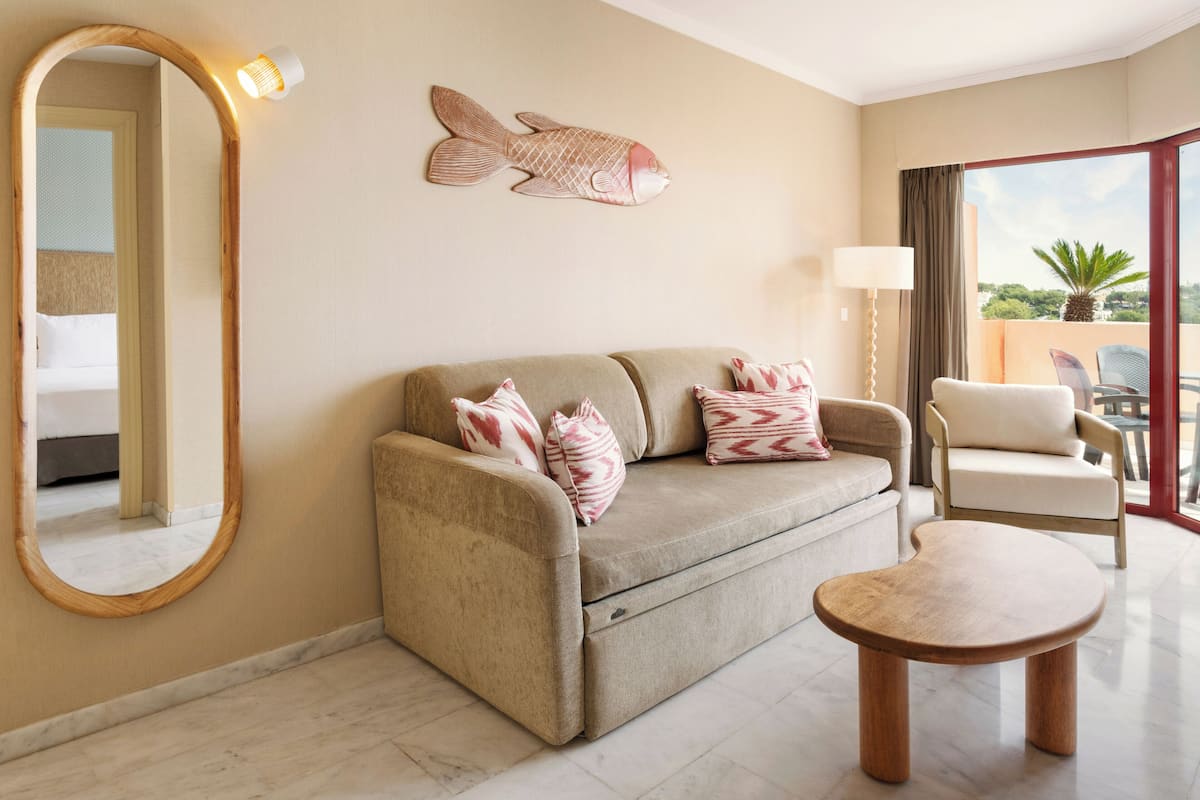 A hotel room with a sofa, a round wooden table, a chair, a fish wall art, a lamp, and a large mirror.