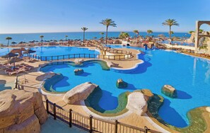 A large outdoor swimming pool with multiple sections and rock formations, surrounded by palm trees and a clear view of the ocean.