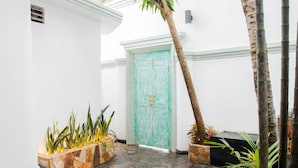 Un camí estret que condueix a una porta turquesa, amb maceta de pedra i plantes tropicals a cada costat.