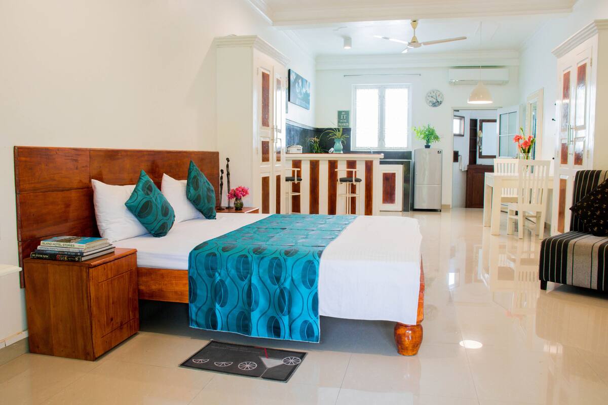 A hotel room with a wooden bed, a small bedside table, a blue patterned bedspread, and a view of the kitchen and dining area.