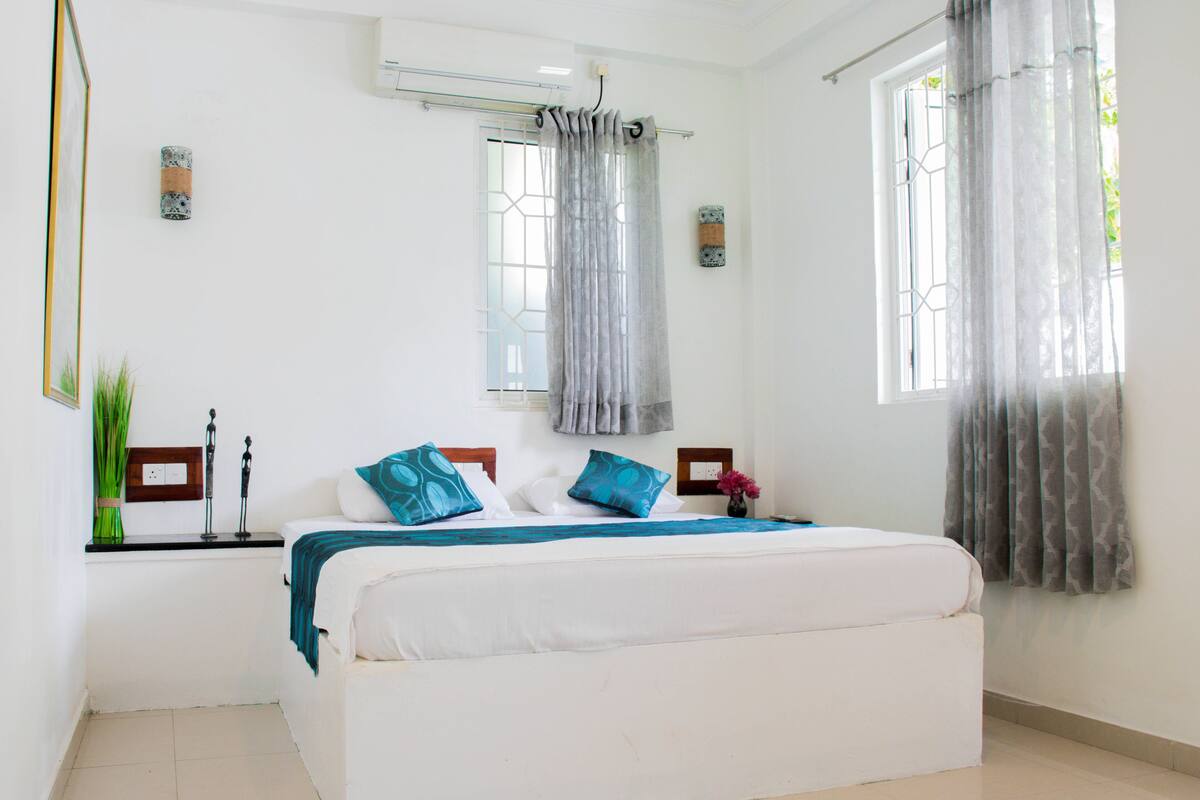 A bedroom with a large bed, a window with curtains, a wall-mounted air conditioner, and a small table with decorative items.