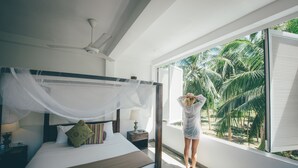 Ein Schlafzimmer mit einem Himmelbett, einem Nachttisch mit Lampe und einem Fenster mit Blick auf Palmen.