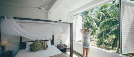 A bedroom with a four-poster bed, a nightstand with a lamp, and a window with a view of palm trees.