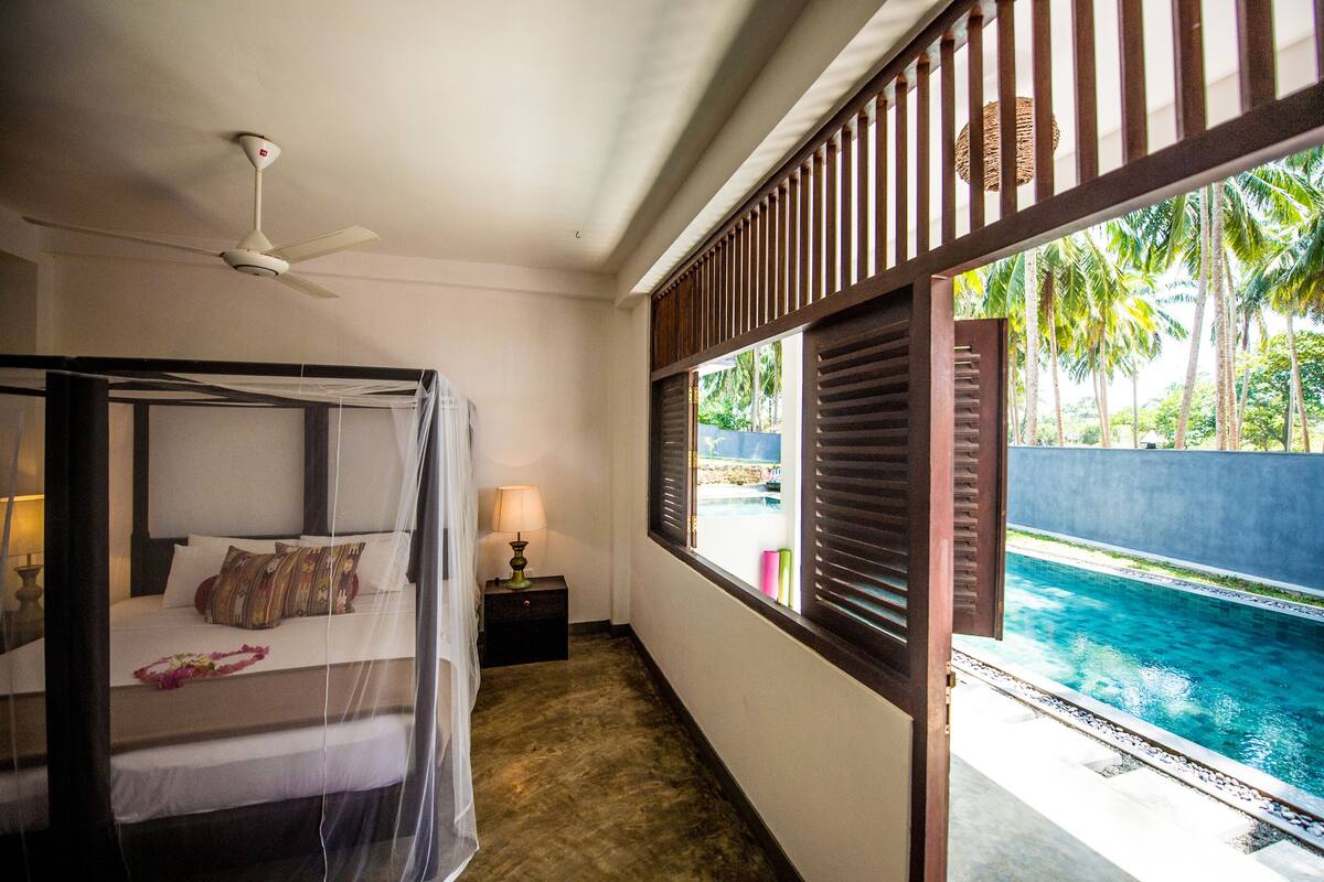 A bedroom with a bed, bedside table, lamp, and a view of a swimming pool.