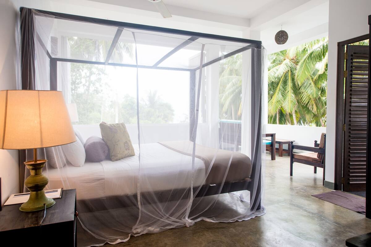 A room with a bed covered by a mosquito net, a bedside lamp, and a view of palm trees through the window.