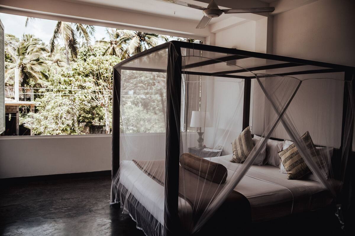 A room with a four-poster bed, a ceiling fan, and a view of palm trees through a large window.