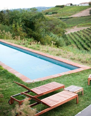 A rectangular swimming pool with a brick border, surrounded by a grassy area with wooden lounge chairs and a small table.