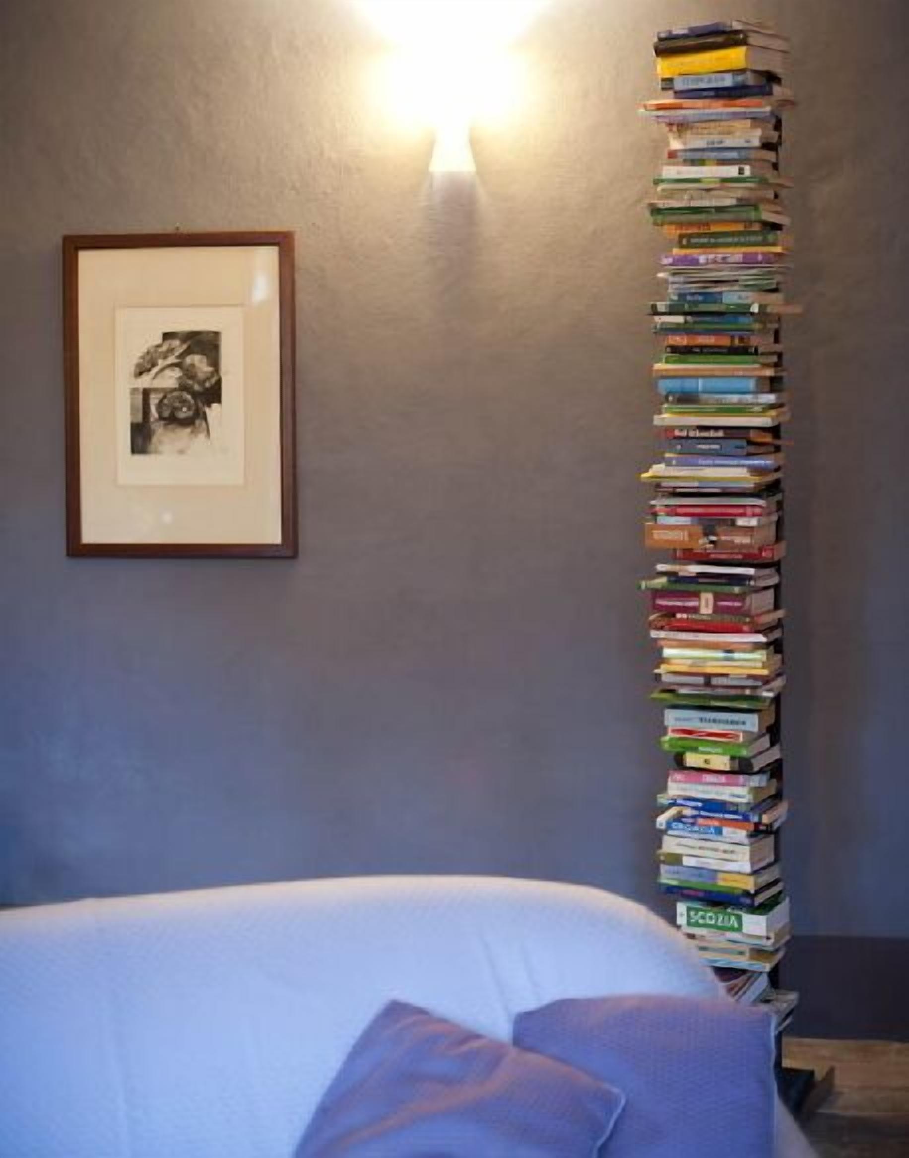 Una pila ordinata di libri appoggiata a una parete, un quadro incorniciato e un letto con un cuscino.