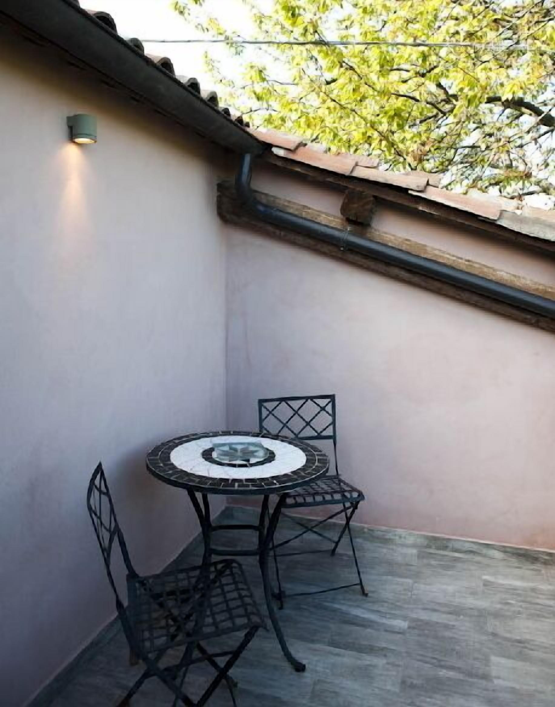 Een klein terras buiten met een ronde tafel, twee stoelen, een wandlamp en een dak met tegels.
