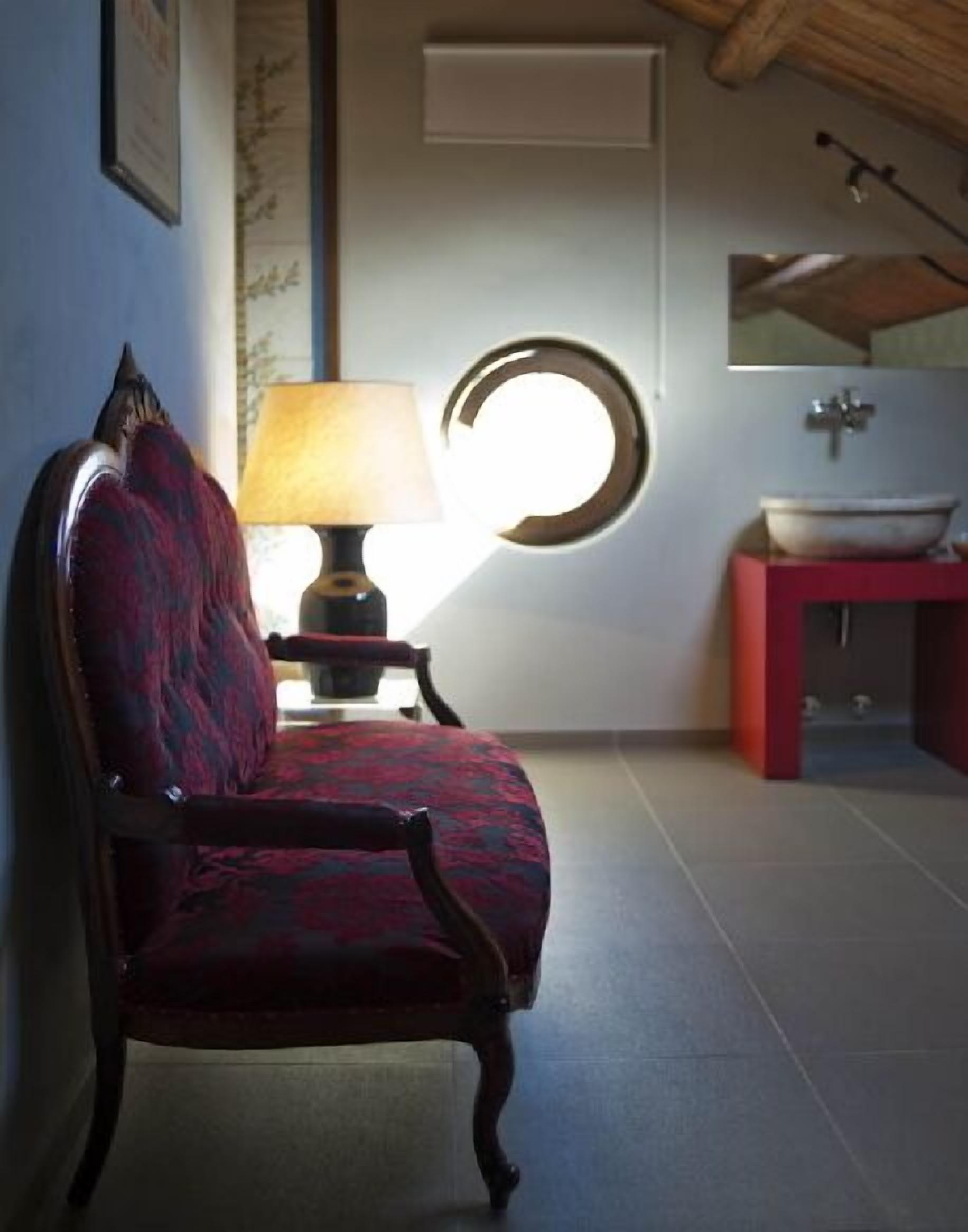 Una silla con un cojín estampado, una lámpara de mesa, un espejo redondo y un mueble rojo con lavabo.