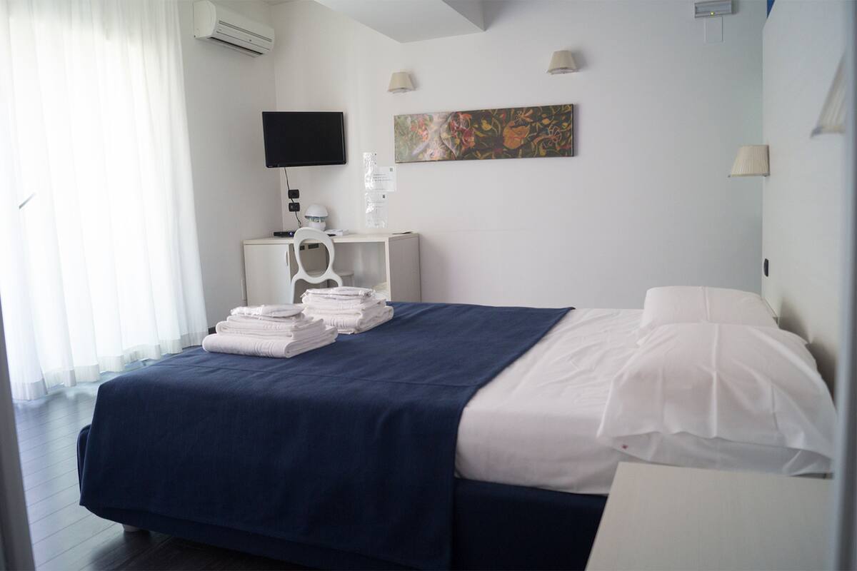 A hotel room with a bed, a desk, a chair, a flat-screen TV, a painting, and a window with curtains.