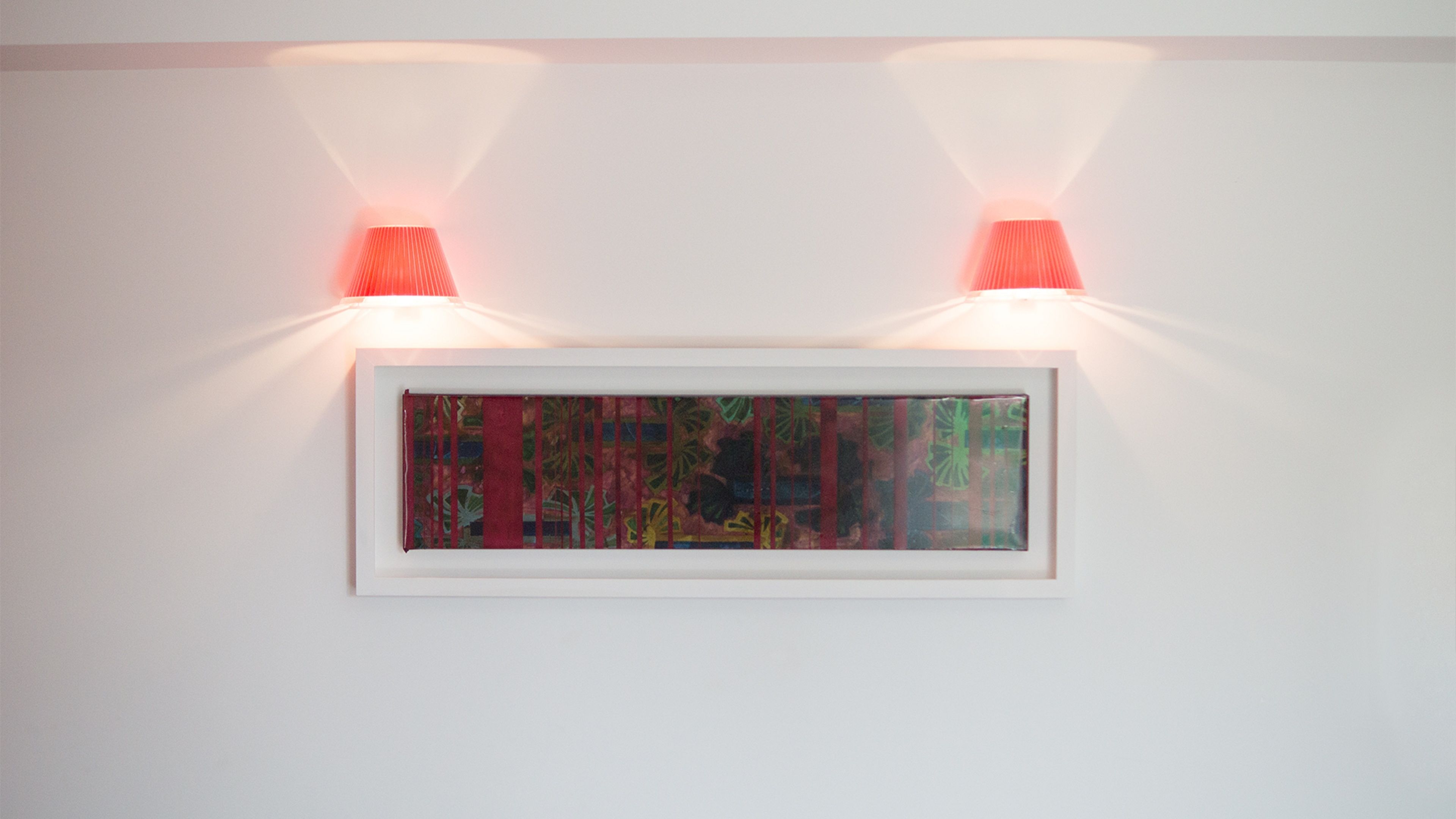 A framed artwork illuminated by two wall-mounted lights on a plain white wall.