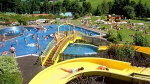 Un espace piscine comprenant plusieurs bassins et un toboggan aquatique, le tout entouré d’une végétation luxuriante et de collines.