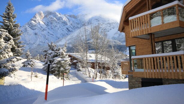 Un chalet en bois doté d’un balcon, entouré d’arbres couverts de neige et avec des montagnes en arrière-plan.