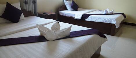 A hotel room with two beds, each with white linens and dark headboards. There is a window with curtains, a bedside table with a remote control, and a decorative pillow on the left bed.