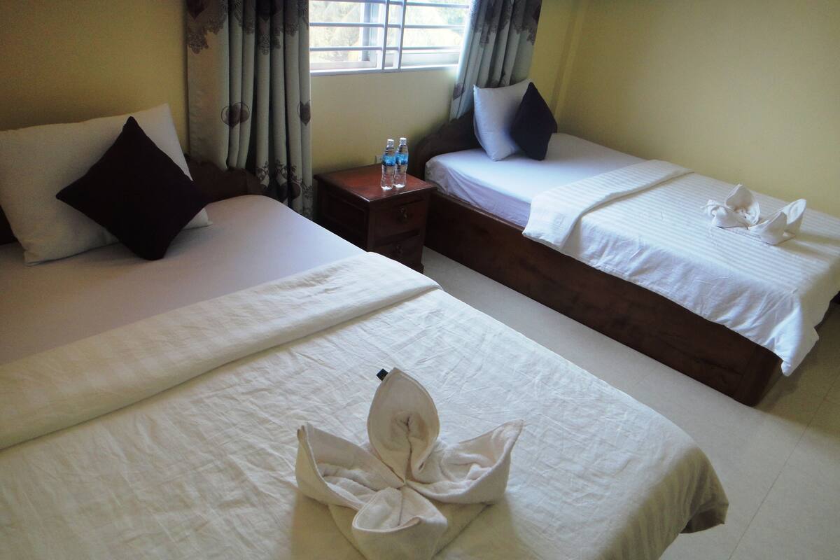 Une chambre d’hôtel avec deux lits, chacun doté de draps et d’oreillers blancs, une petite table de chevet en bois avec des bouteilles d’eau, et une fenêtre avec des rideaux.