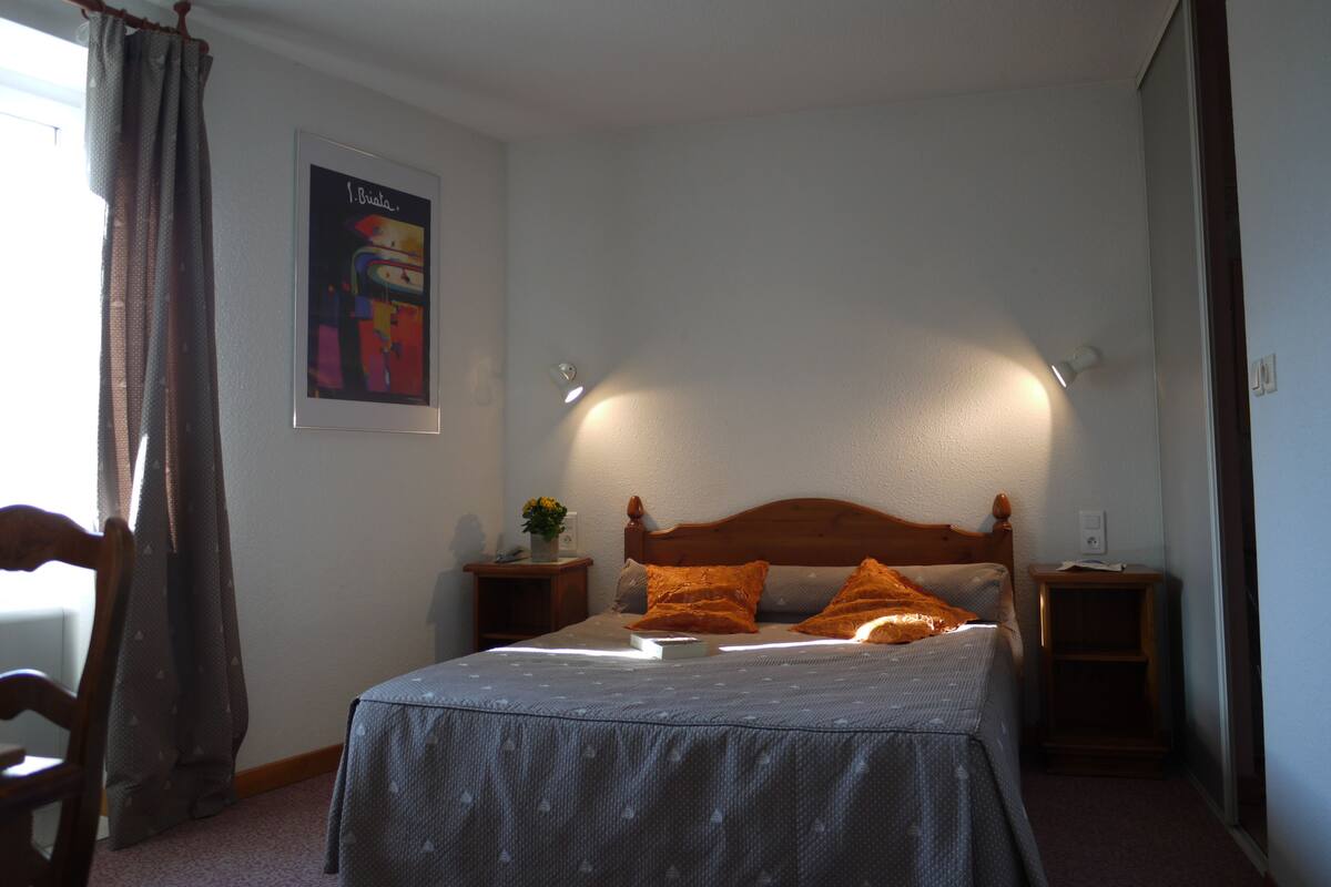 Un dormitorio con cama, cabecero de madera, dos mesitas de noche, una pequeña mesa con una planta y un póster en la pared.