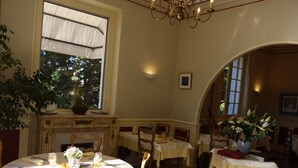 Un espace repas avec des tables rondes, des chaises et une cheminée. De grandes fenêtres laissent entrer la lumière naturelle et un lustre est suspendu au plafond.