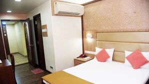 A hotel room with a large bed, a bedside table, a nightstand, an air conditioning unit, a wall-mounted lamp, and a door leading to another room. - Hotel Indiana Inn (Navi Mumbai)