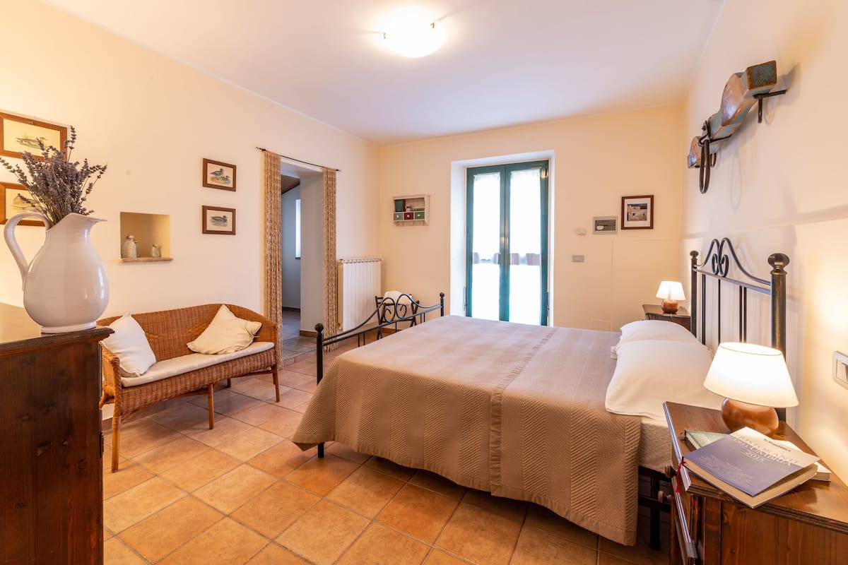 A bedroom with a bed, a nightstand, a lamp, a wicker sofa, a vase with flowers, and a door leading to another room.
