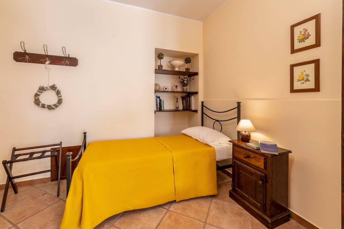 A bedroom with a bed, a nightstand, a shelf with books and decorative items, a coat rack, and a wall-mounted shelf with a bowl and flowers.