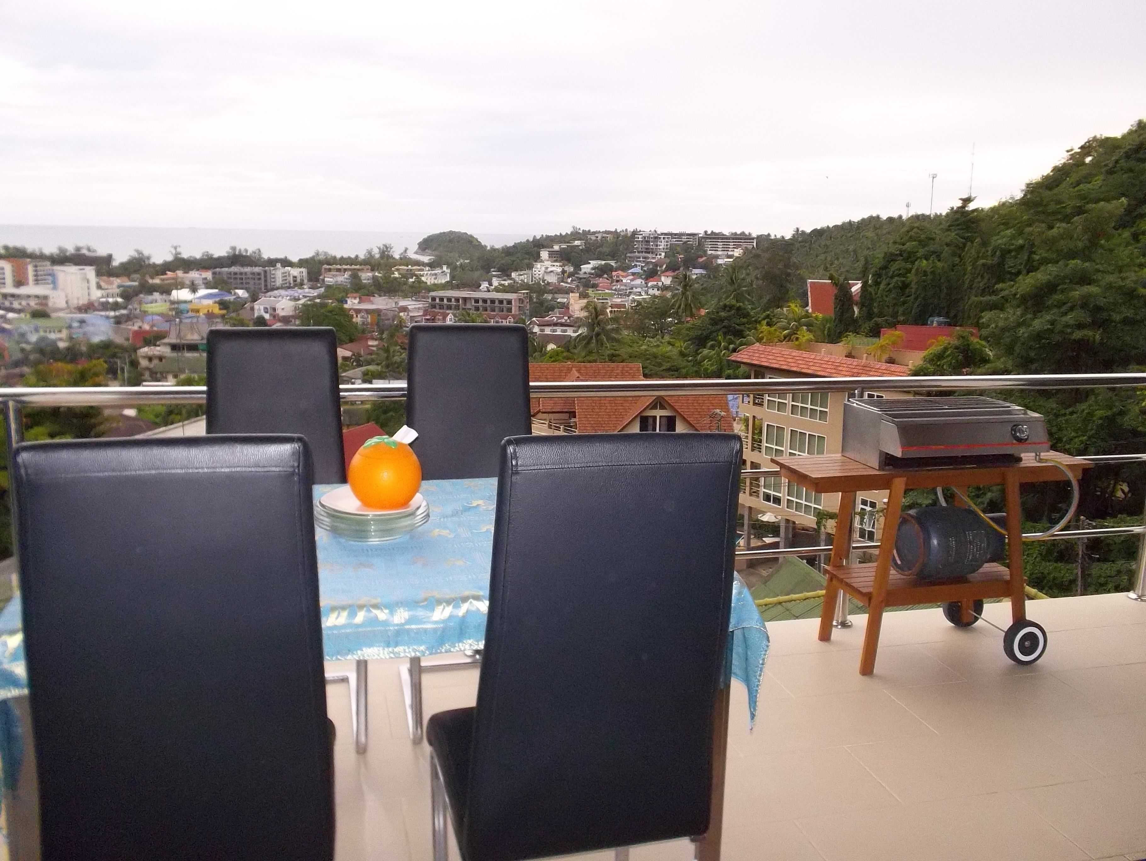 A balcony with a table set for two, overlooking a cityscape with buildings and a view of the sea.