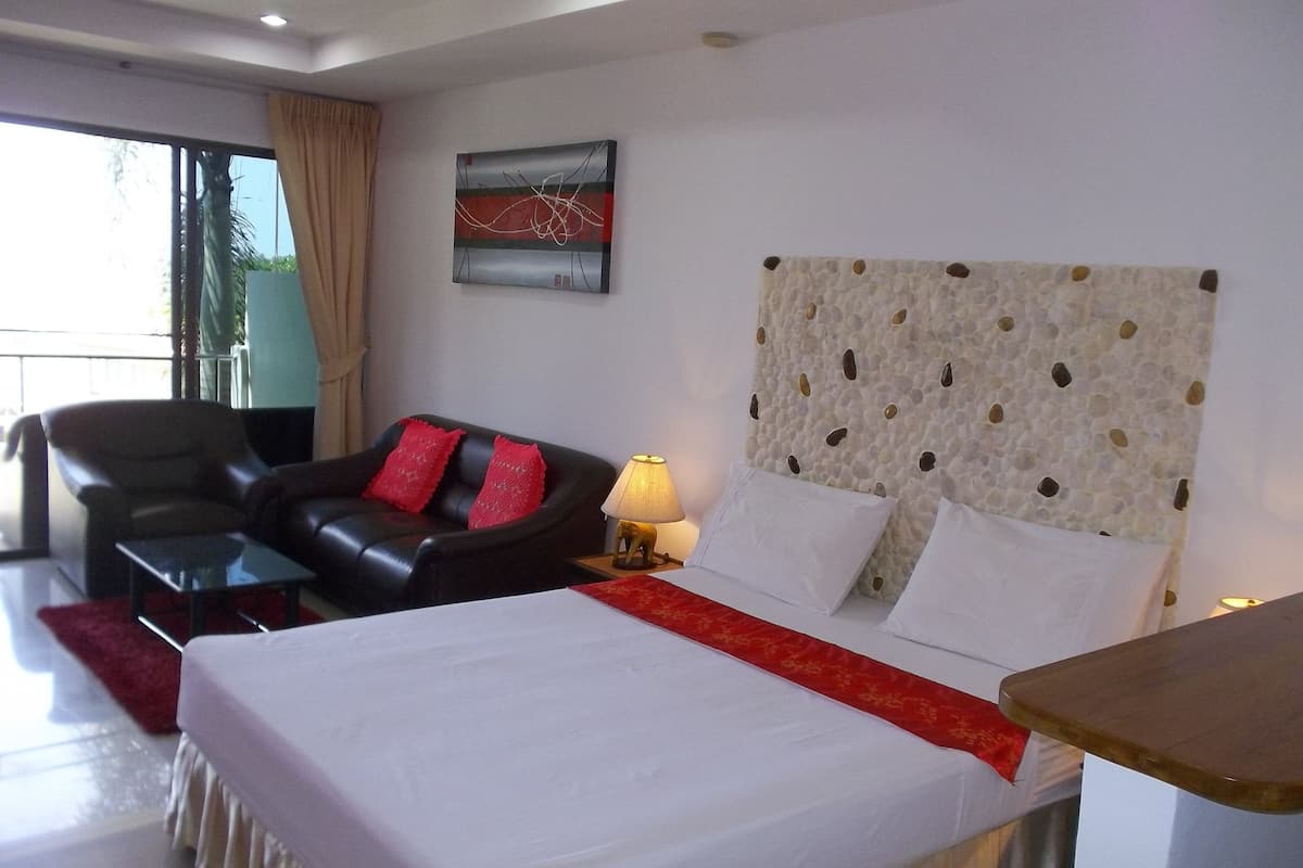 Quarto de hotel com cama, sofá de couro preto com almofadas vermelhas, mesa pequena, abajur e parede com cabeceira texturizada.