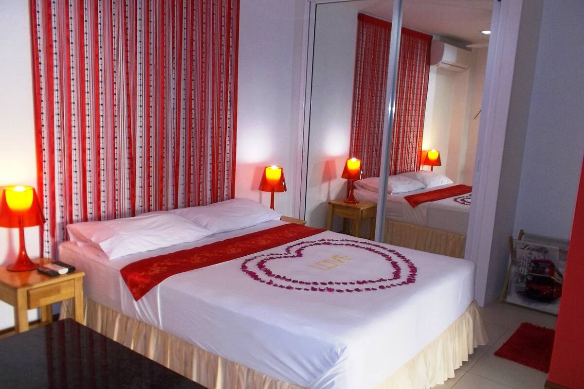 Quarto de hotel com uma cama, duas luminárias laterais, um espelho e cortina listrada em vermelho e branco.