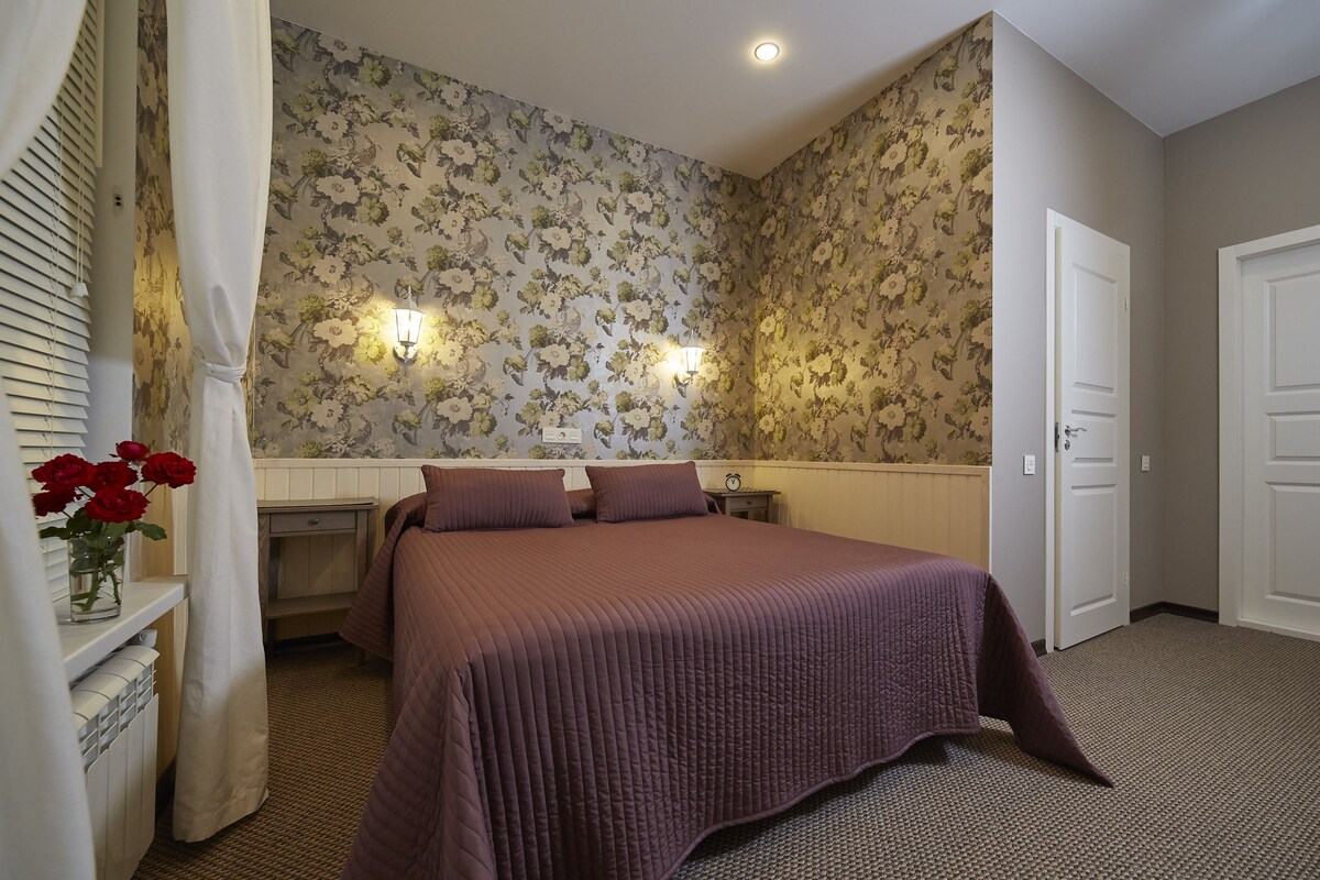 A bedroom with a large bed, floral wallpaper, two wall-mounted lights, and a door.