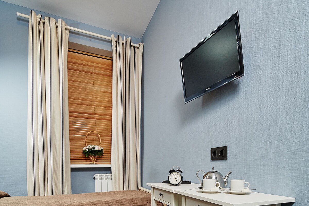A bedroom with a bed, a bedside table with a clock and teapot, a TV mounted on the wall, and a window with curtains.