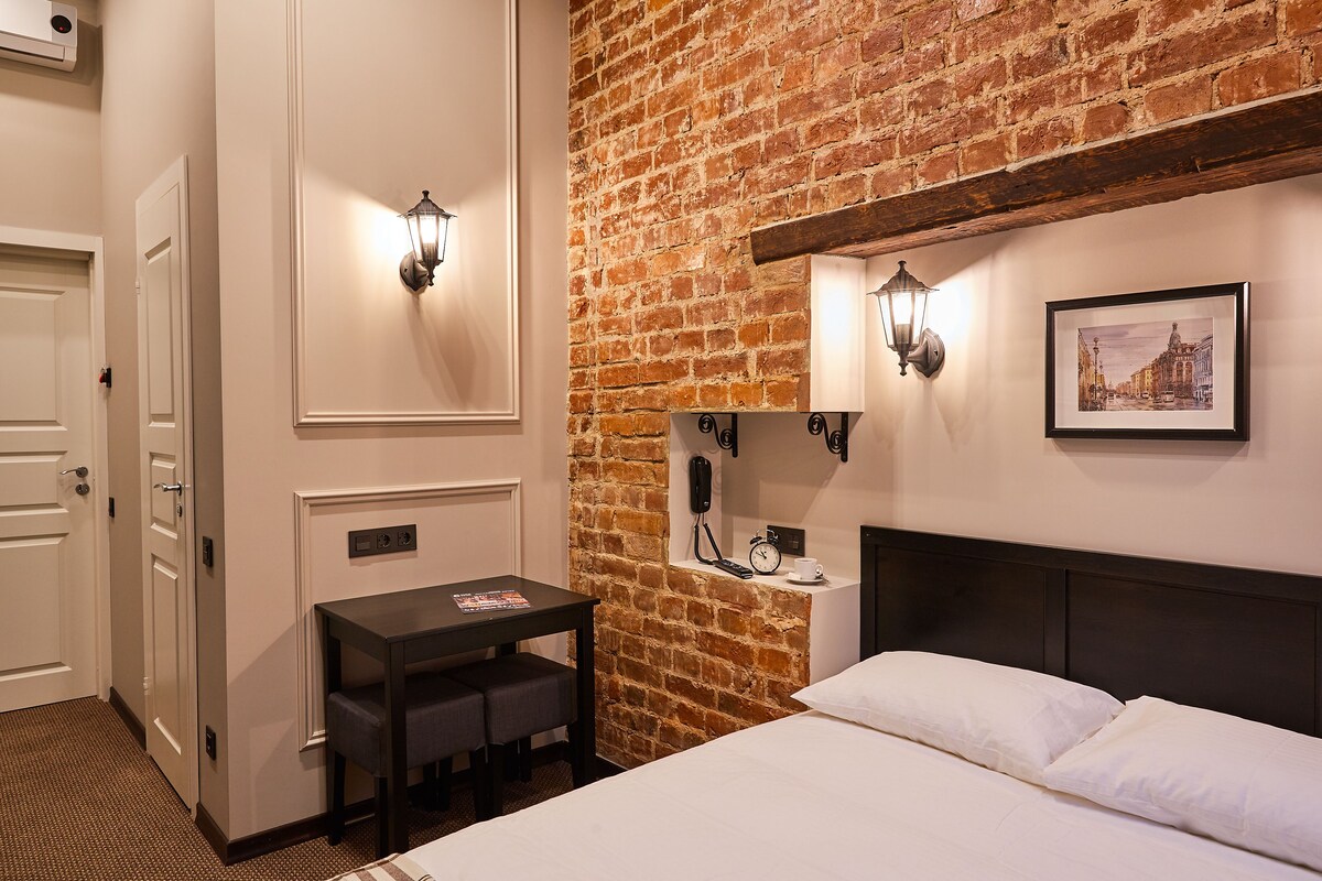 A hotel room with a brick wall, a bed with white linens, a small desk with a chair, and two wall-mounted lamps.