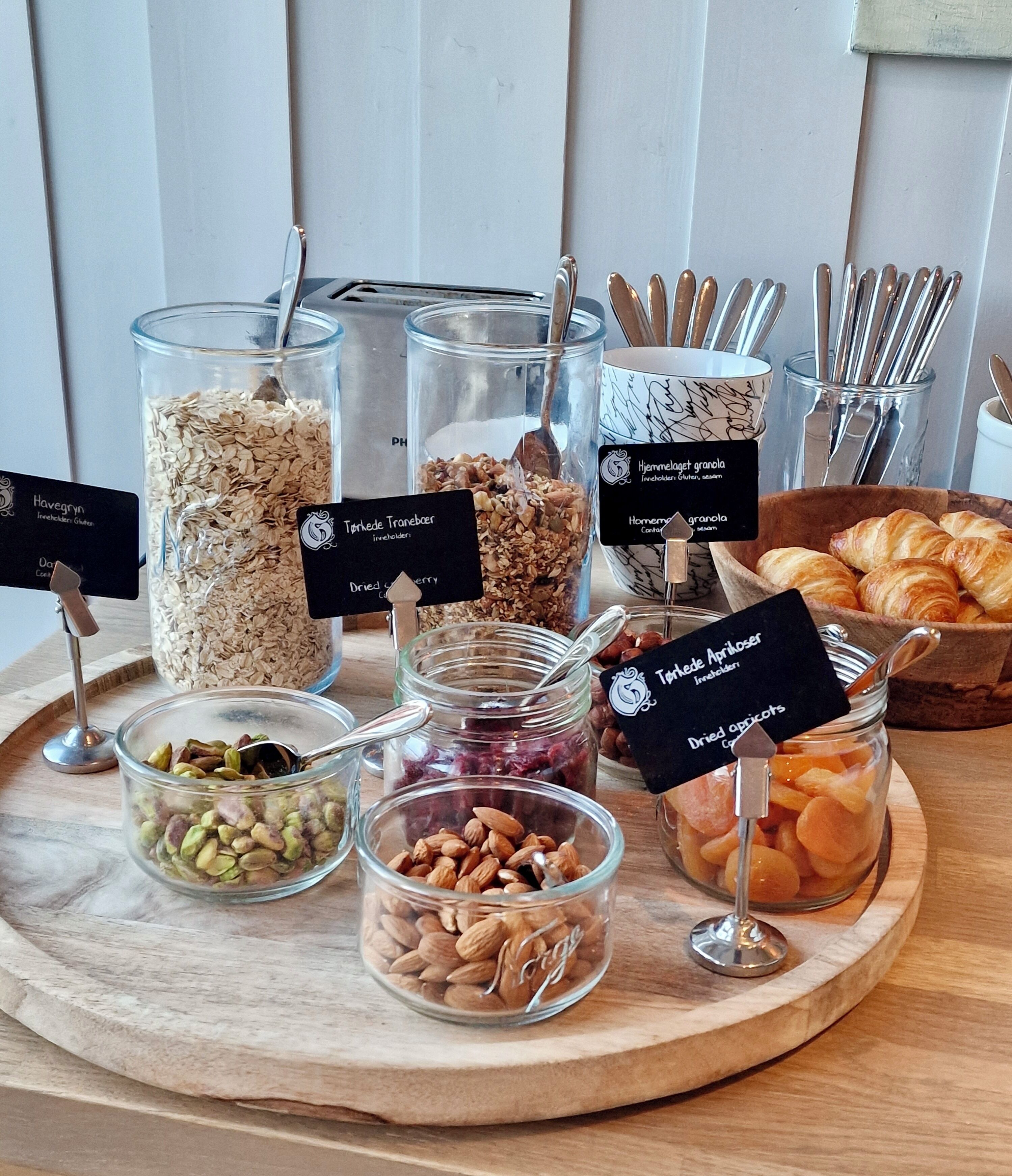 Variedad de alimentos sobre una tabla de madera, como granos, nueces, frutas secas y pasteles, cada uno con su nombre.
