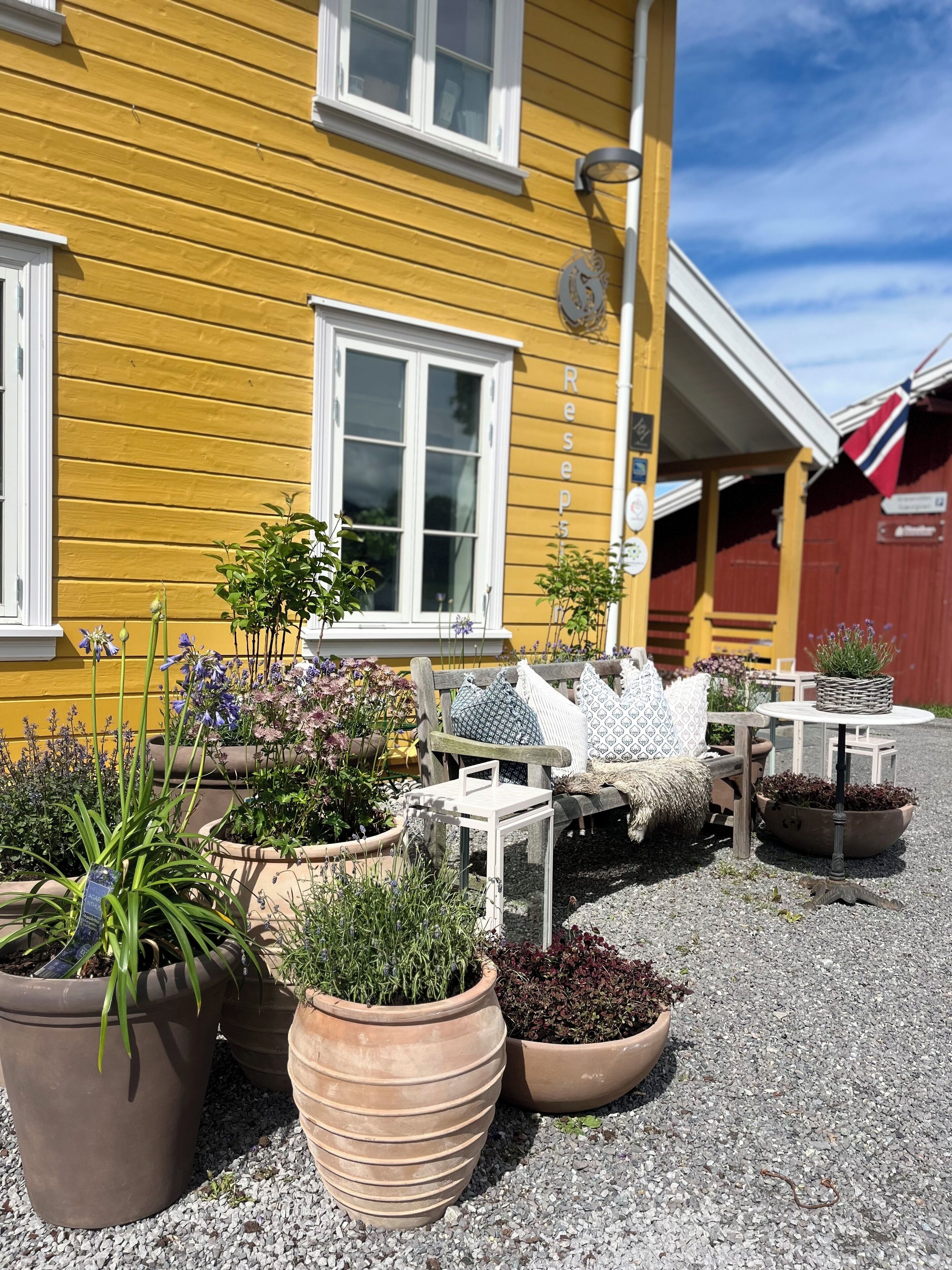 A yellow building with a window and a door, surrounded by potted plants and outdoor furniture.