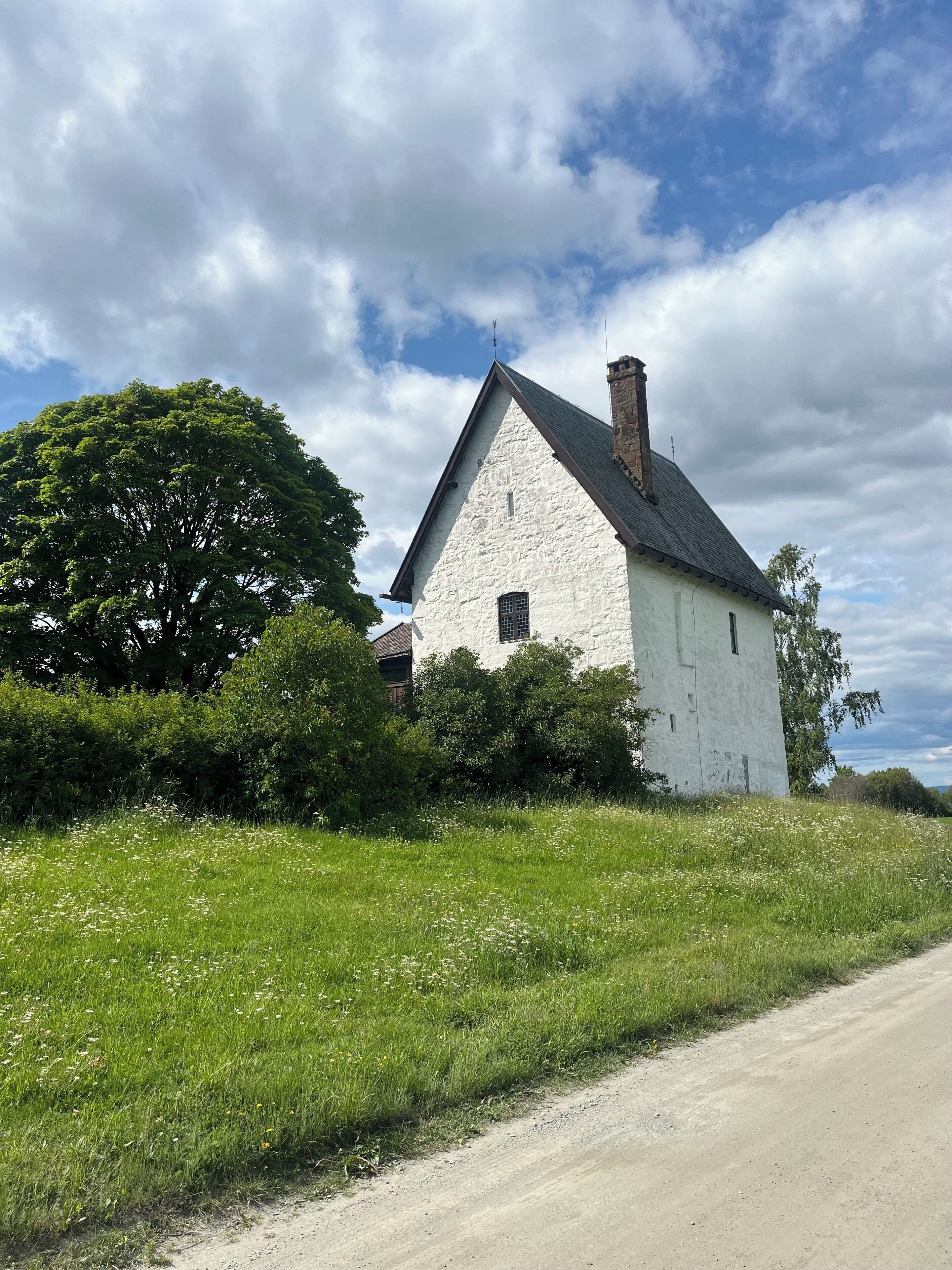 A white stone house with a steeply pitched roof, surrounded by lush greenery and a clear sky.