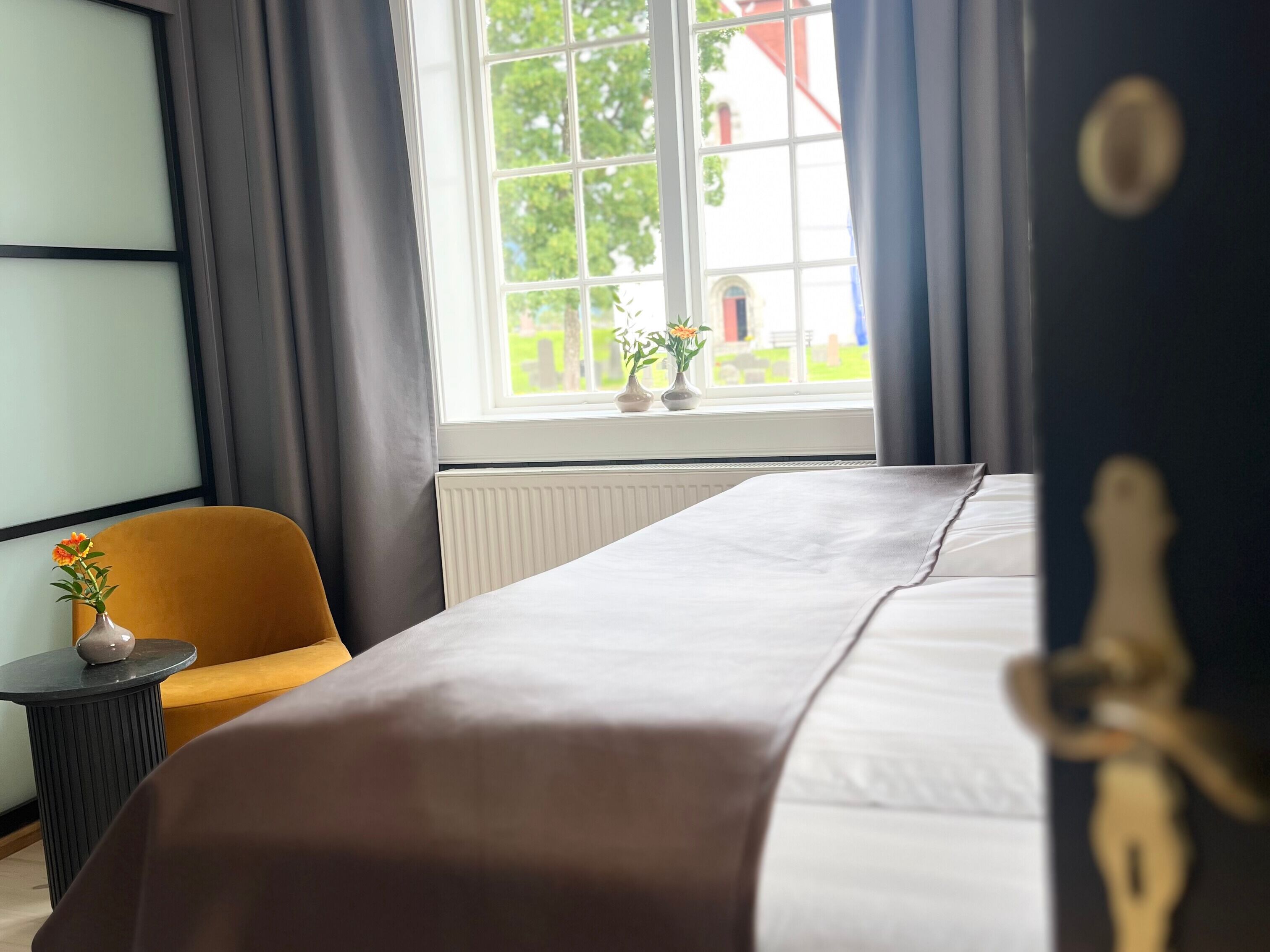 A hotel room with a bed, a small table with a vase of flowers, a chair, and a window with curtains.